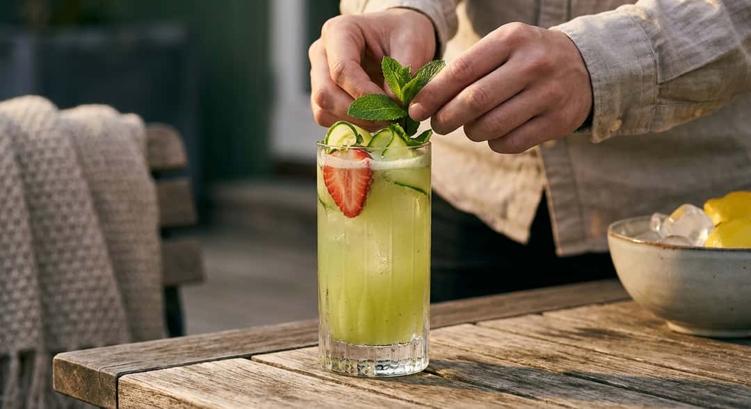Large glass with a light-green drink, topped with strawberries, cucumber slices, and a mint sprig, standing on a wooden outdoor panel in soft spring light
