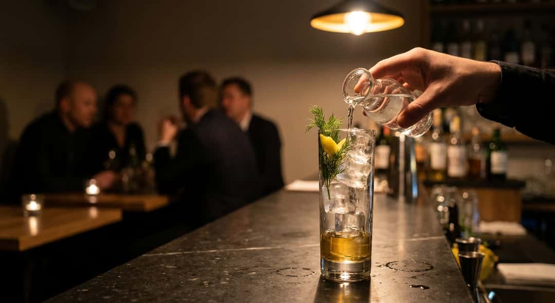 Nærbilde av en Akevitt Tonic under tilberedning – tonic water helles forsiktig i et høyt glass fullt av is, dill og sitr