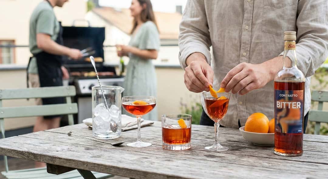 Kolme erilaista aperitiivilasia katkerolikööricocktaileilla ja koristeluilla ulkona puupöydällä, taustalla grillaileva väkijoukko