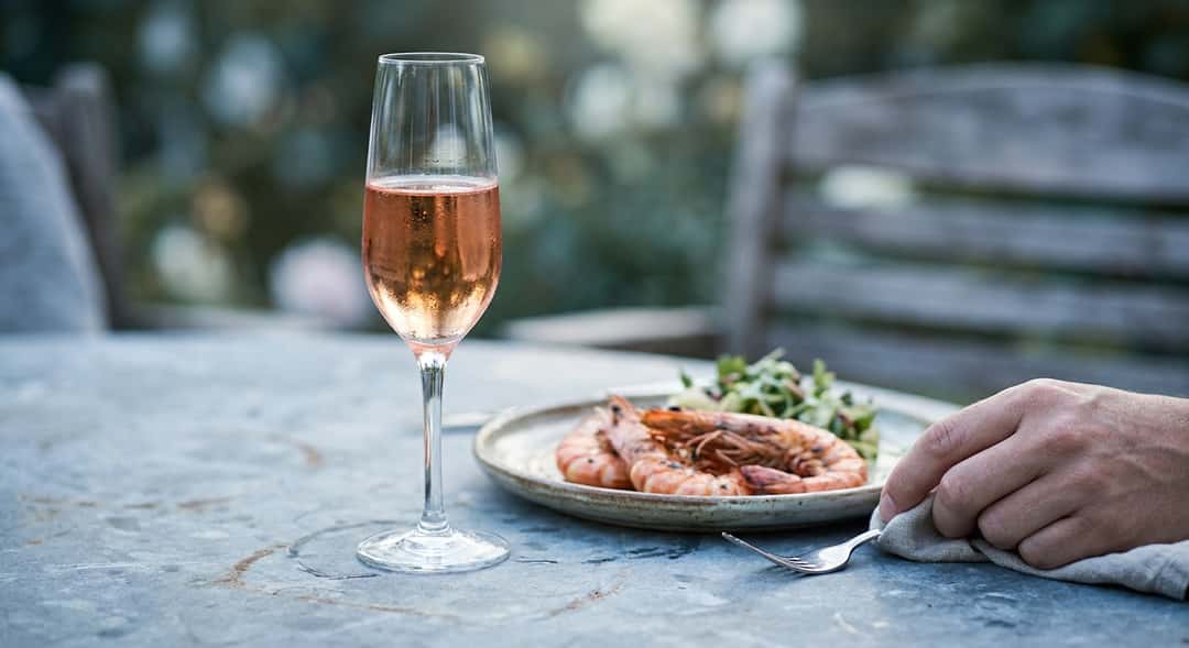 Glass of cava rosado in a champagne flute, salmon colour, grilled prawns on a plate alongside, summer evening outdoors