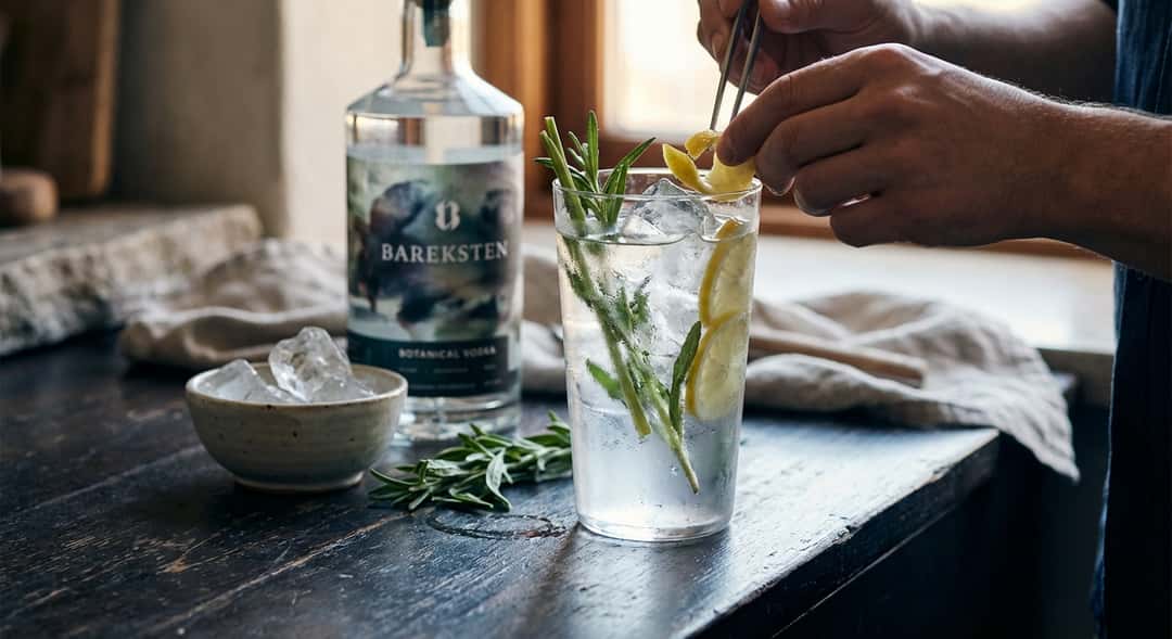 Highballglass med klar drink, frodige grønne urtestilker, isbiter og sitronskiver, dugget glass på trebord, (Bareksten B