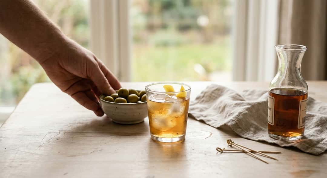 A glass of dry sherry with ice and a bowl of green olives on a simple wooden table in natural evening light