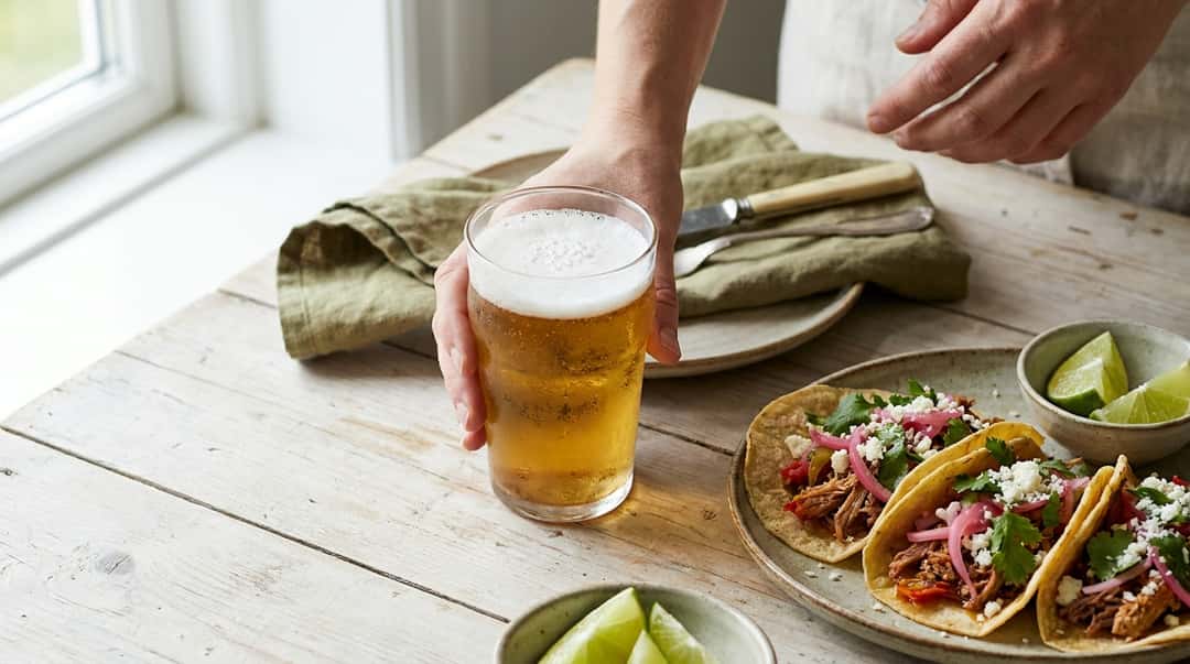 Et kaldt glass lagerøl med kondens på utsiden, servert ved siden av en tallerken med fargerike tacos og limebåter
