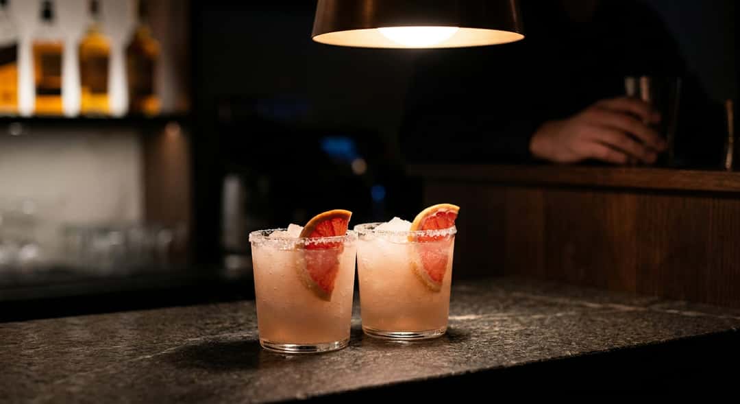 To frosne Paloma-glass med grapefruktskiver og salt rim, servert på en hvit treterrasse i sommersol