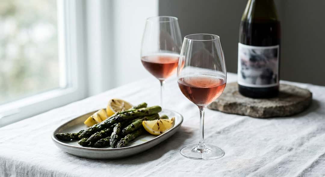 Two glasses of light red wine on a white set table with grilled asparagus and lemon wedges, natural daylight from a window