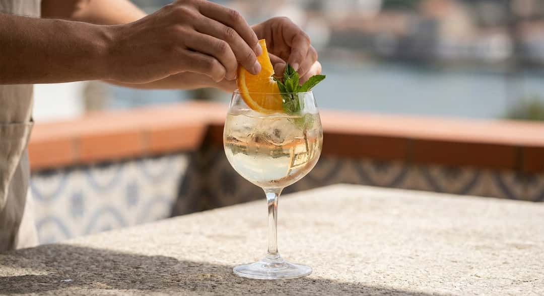 Et stort glass hvit port tonic med is, appelsinskive og mynte på en solrik portugisisk terrasse ved Douro-elven