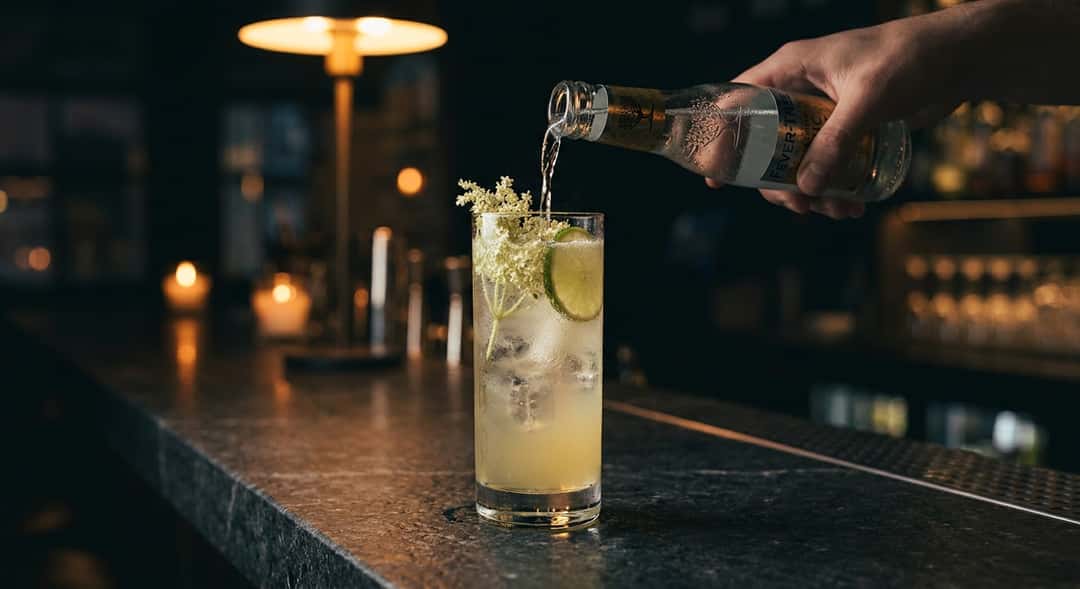 Nærbilde av hylleblomst-smash i et høyt glass med is, friske hylleblomstkvister som pynt og tonic water som helles over,