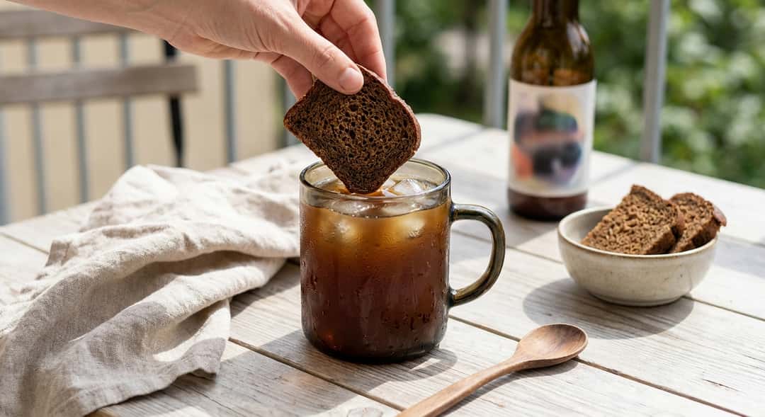 Et mørkt, kondenserende glass med brun kvass på et trebord ute i sommervarmen, isbiter og rugbrødskive som garnityr