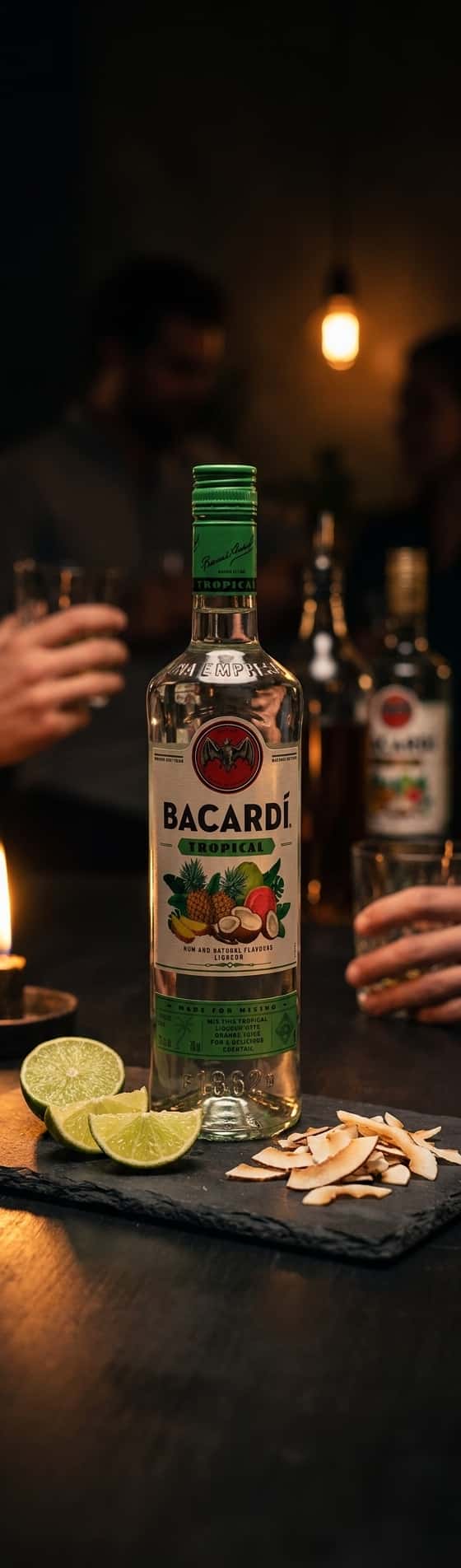 En flaska Bacardi Tropical på en träbricka bredvid färska limefrukter och kokosflarn, dagsljus från sidan