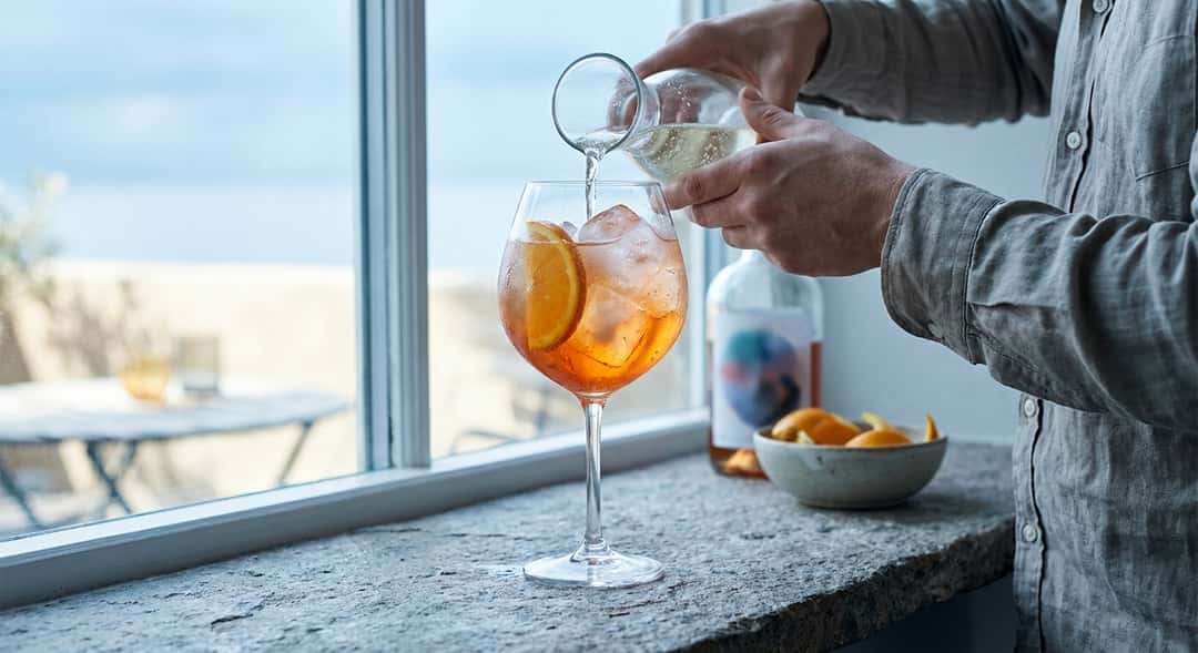 A lush spritz in a large balloon glass filled with ice and an orange slice, sunshine and blue sky in the background (Aperol)