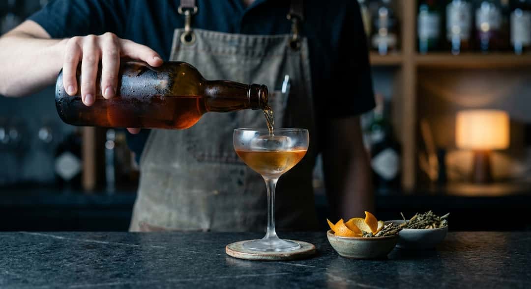 Ung bartender på en moderne bar som heller iskald Madeira i et elegant glass, mørk bakgrunn med stemningsfullt lys
