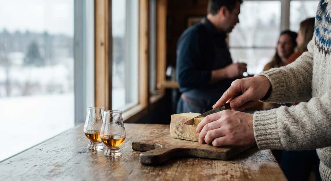 Hender som skjærer ost på trevare, små glass med brennevin og lys vinterkvelds-atmosfære rundt, hygge og ro