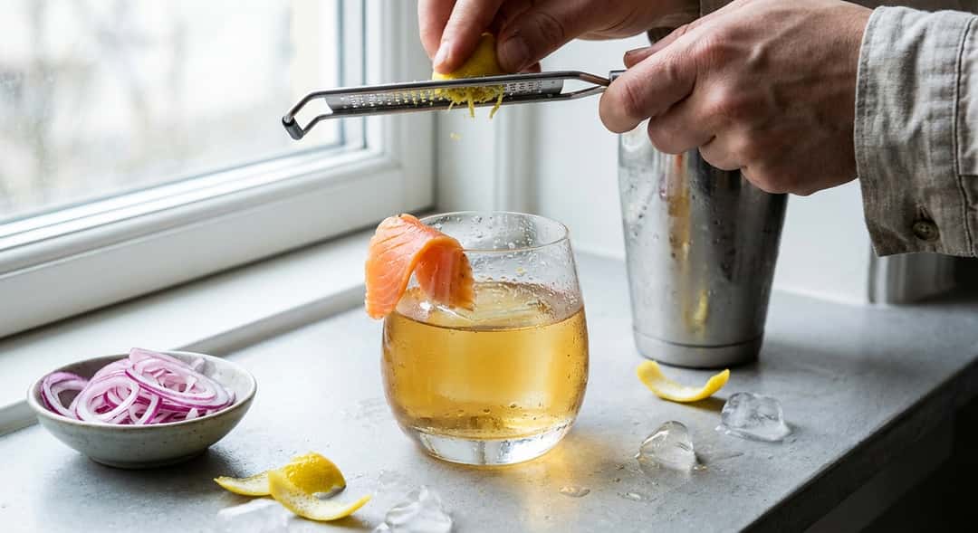 Närbild av akvavitdrink med tunn laxskiva draperad över glaskanten, droppar kondenserar på glaset, citronskal