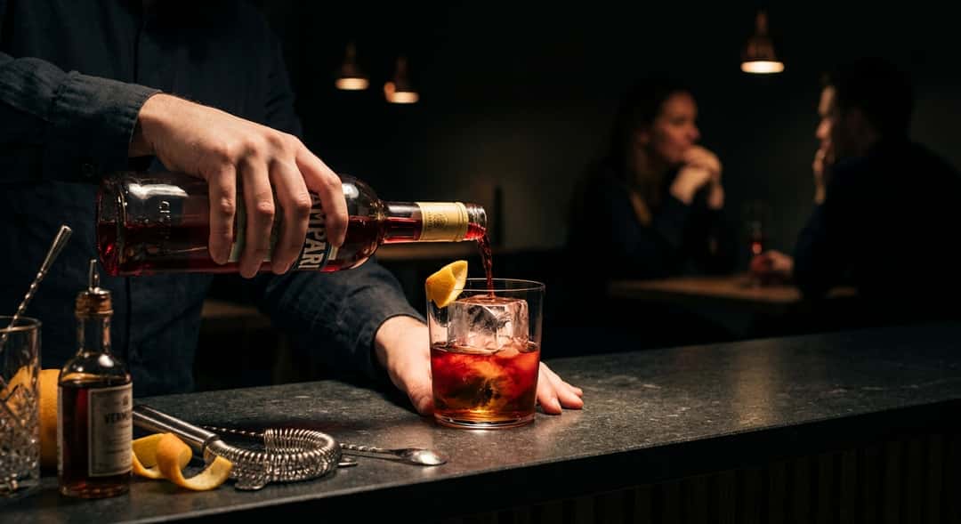 Lähikuva Camparistä, jota kaadetaan jään päälle rocks-lasiin, punainen väri leviää alaspäin (Campari Bitter)