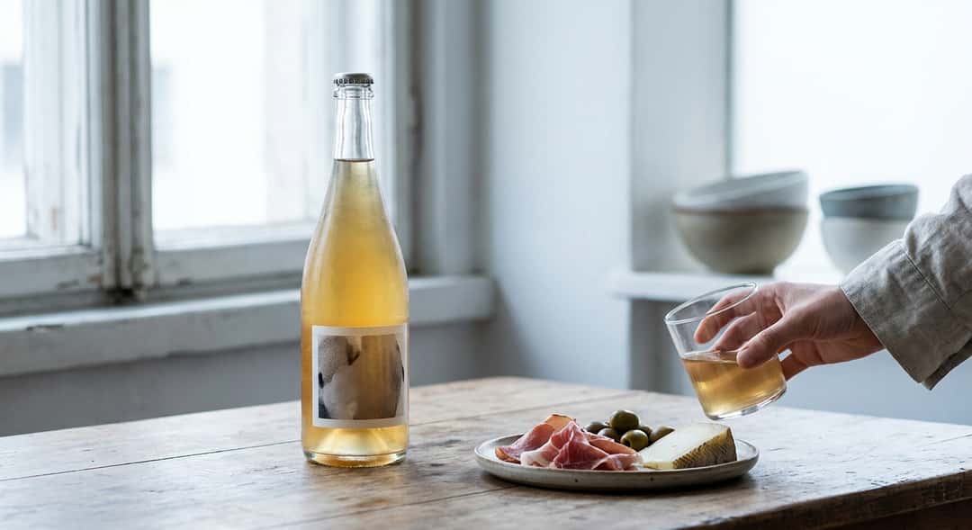 En flaska pét-nat på ett träbord bredvid ett fat med tapas och färsk ost, ljust och luftigt stämningsbild