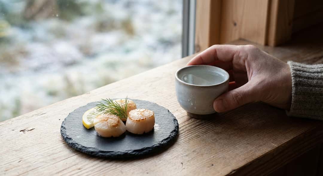 Färska norska kammusslor på skiffer med ett glas sake bredvid, nordisk bordsdukning i dagsljus