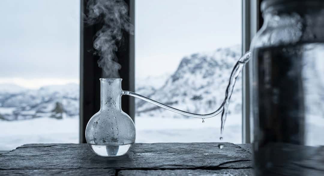A distillation flask with steam rising, a backdrop of snow-covered mountains, water dripping from a pure source into the production process