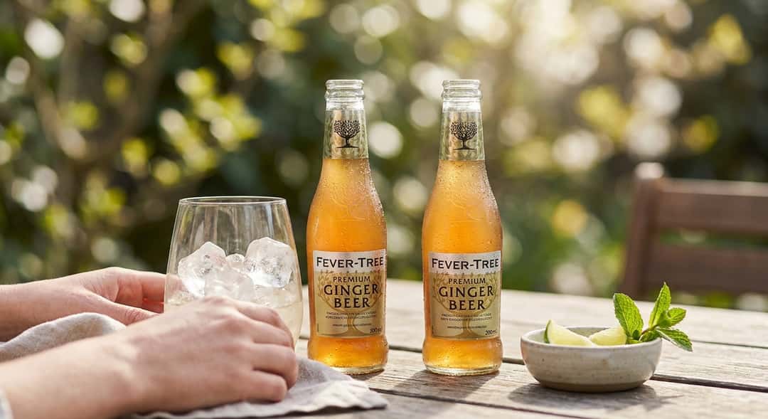 To flasker ginger beer på en trebord ute på terrasse, vårgrønt i bakgrunnen, naturlig dagslys