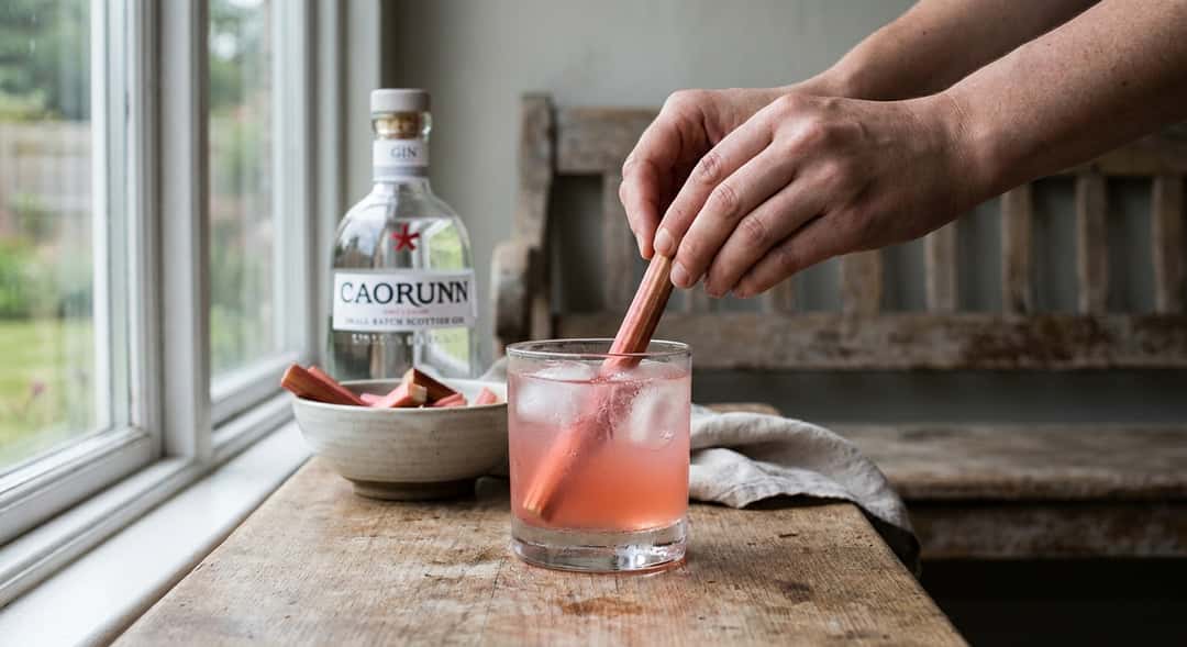 Rabarbra-fizz i rocks-glass med isbiter og rå rabarbrabit som garnityr, rustikk trebenk i bakgrunnen (Balmenach Caorunn
