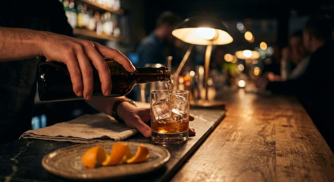 Närbild av vermouth som hälls över stora isbitar i ett lågt glas, med ett apelsinskal i förgrunden